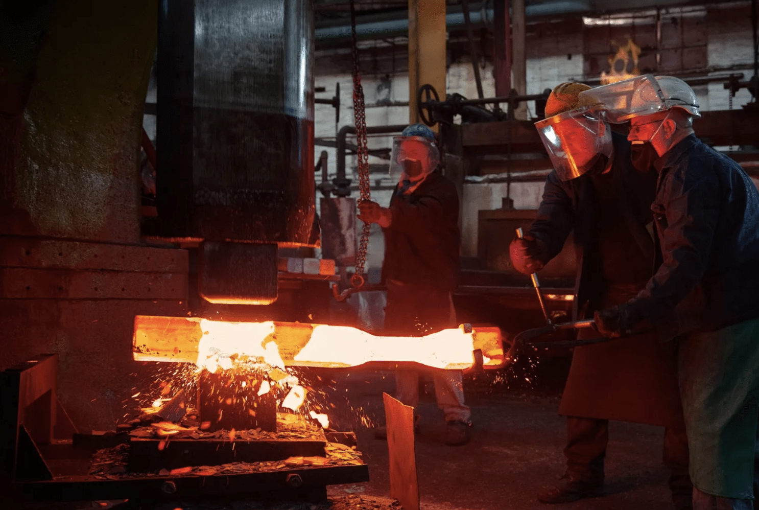 workers using power hammer for forging, industrial workers handling heated metal in a factory